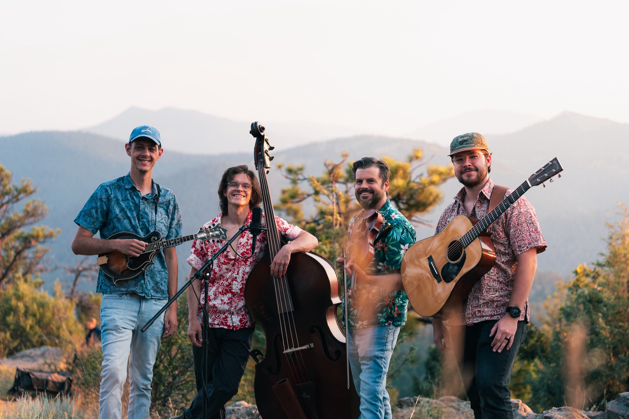 The four Bikini Boys in Hawaiian shirts on a foothills ridge at sunset, holding a mandolin, upright bass and guitar.